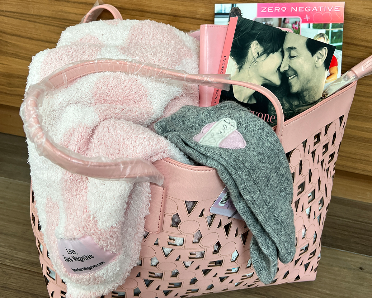 Pink Chemo Love Tote on wooden floor, filled with cozy socks, fuzzy blanket, and Zero Negative magazine featuring a couple embracing.