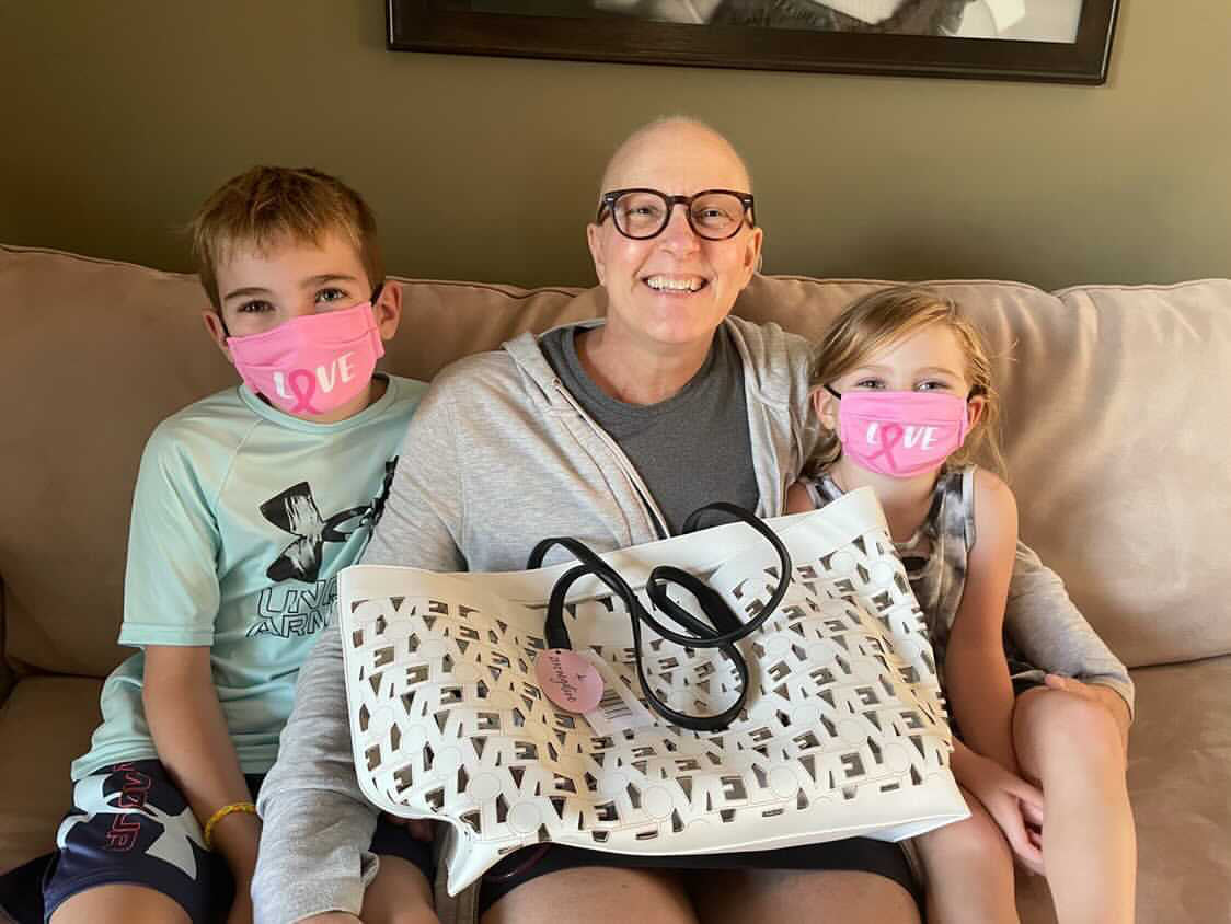 Smiling woman with a bald head, sitting on a couch with two children, all wearing pink “LOVE” masks and holding a white Chemo Love Tote from Zero Negative Foundation.
